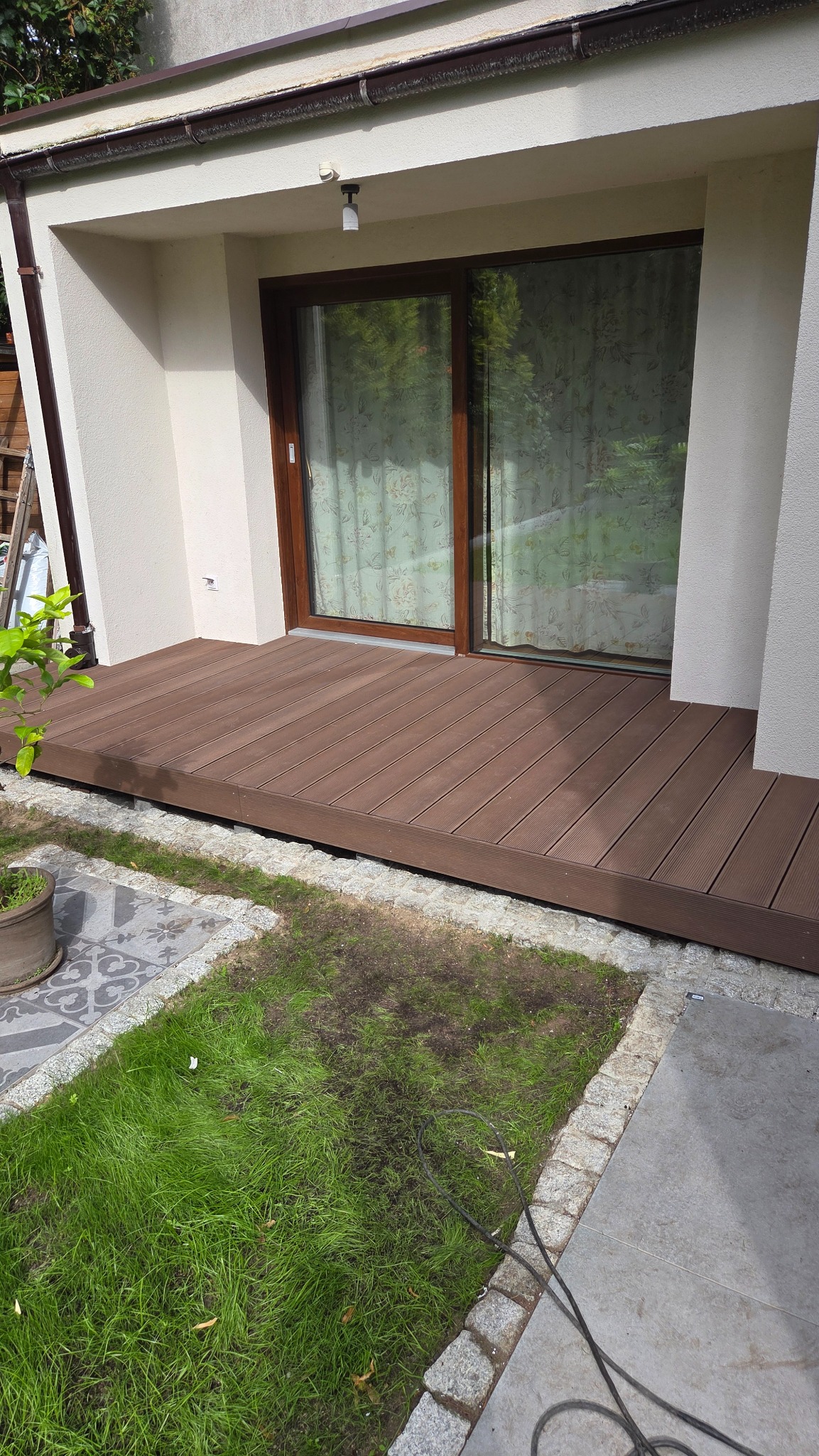 Gotowy taras kompozytowy przylegający do domu, z widocznymi deskami ułożonymi pod kątem, graniczący z trawnikiem i płytami chodnikowymi.