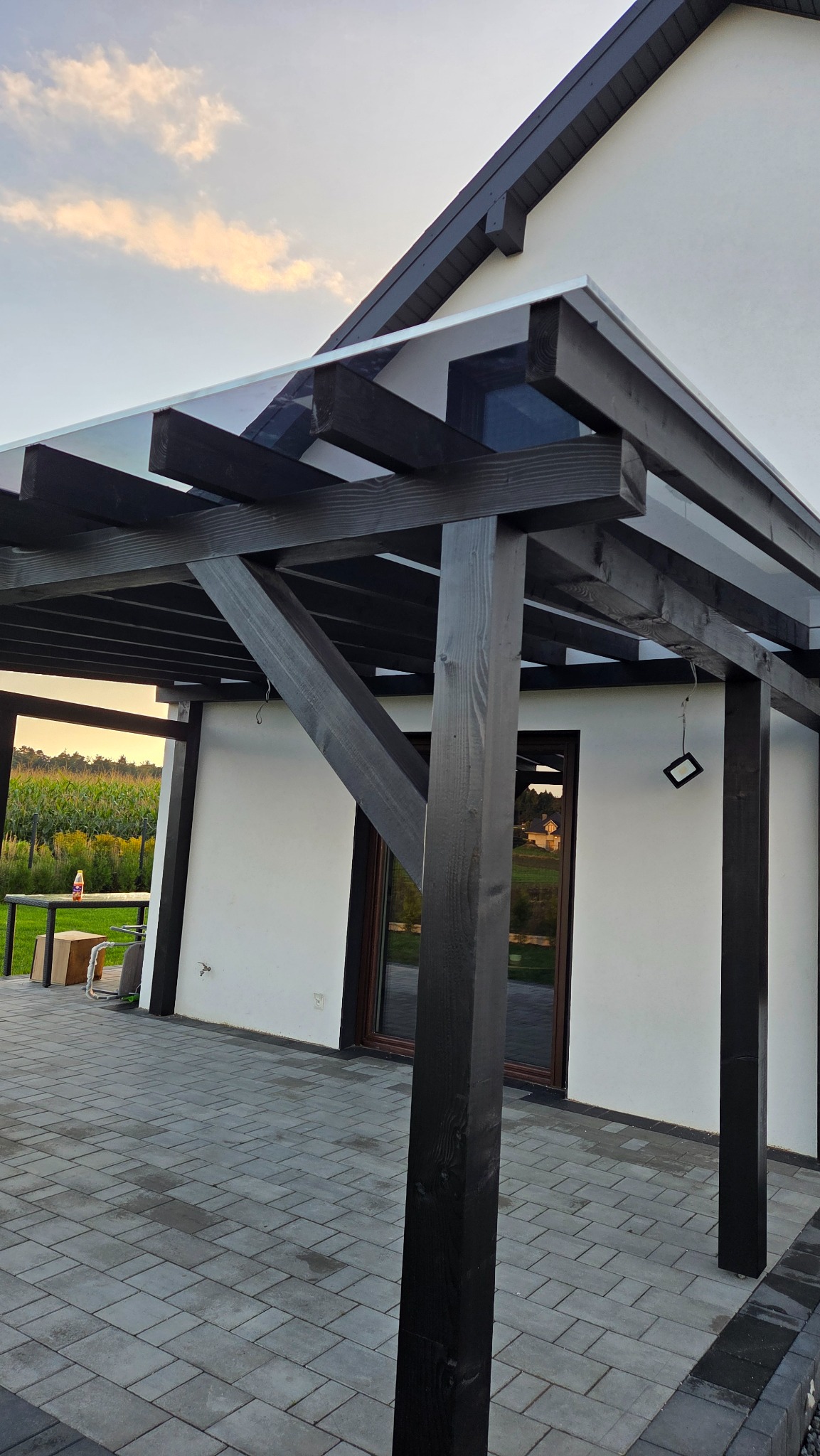 Nowoczesna pergola z ciemnego drewna i przezroczystym zadaszeniem, przylegająca do białego budynku. Widok na ogród w tle, kostka brukowa na ziemi.