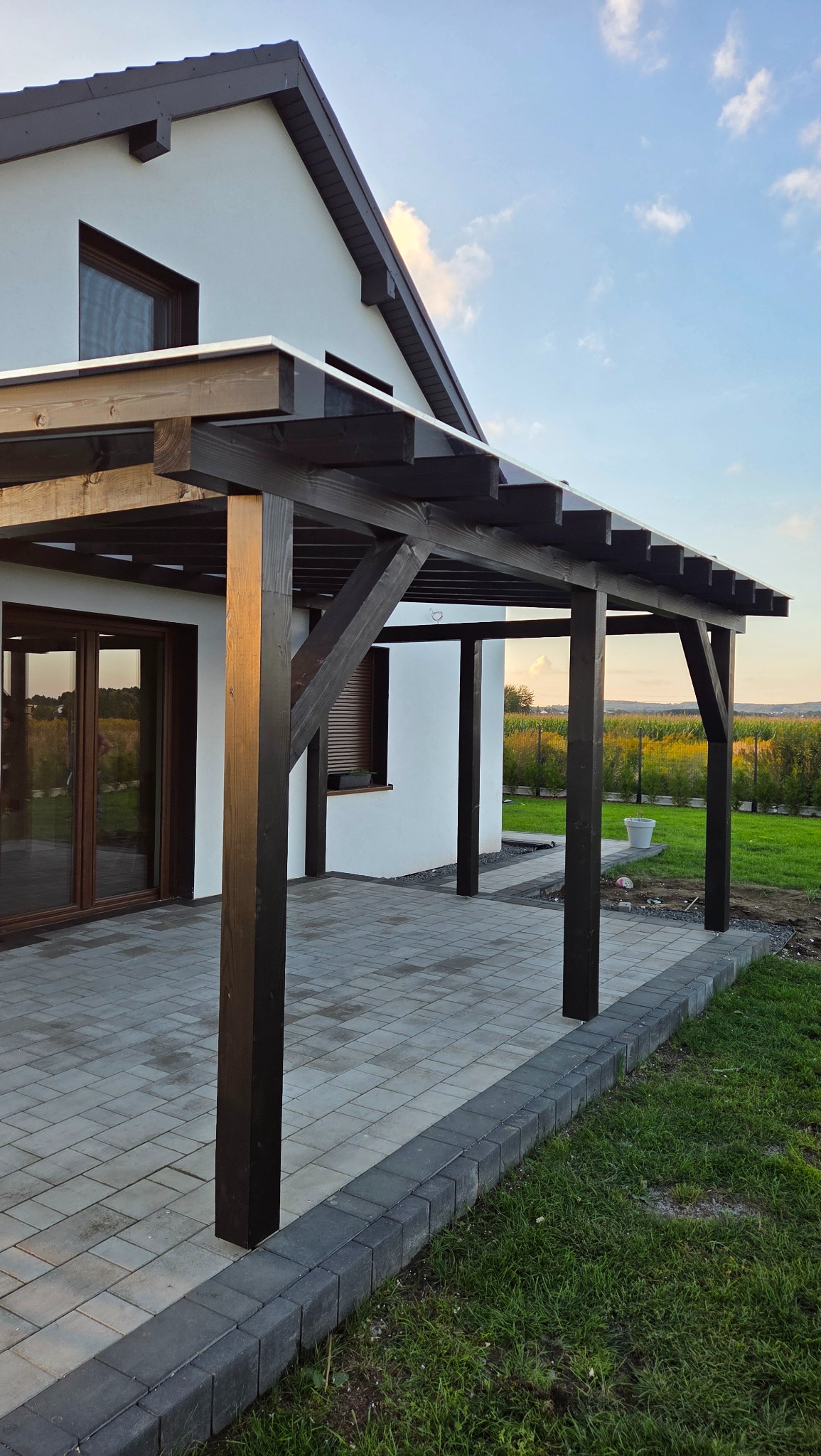 Nowoczesna, brązowa pergola z zadaszeniem przy białym domu, widok z boku na taras z kostki brukowej i zielony trawnik.