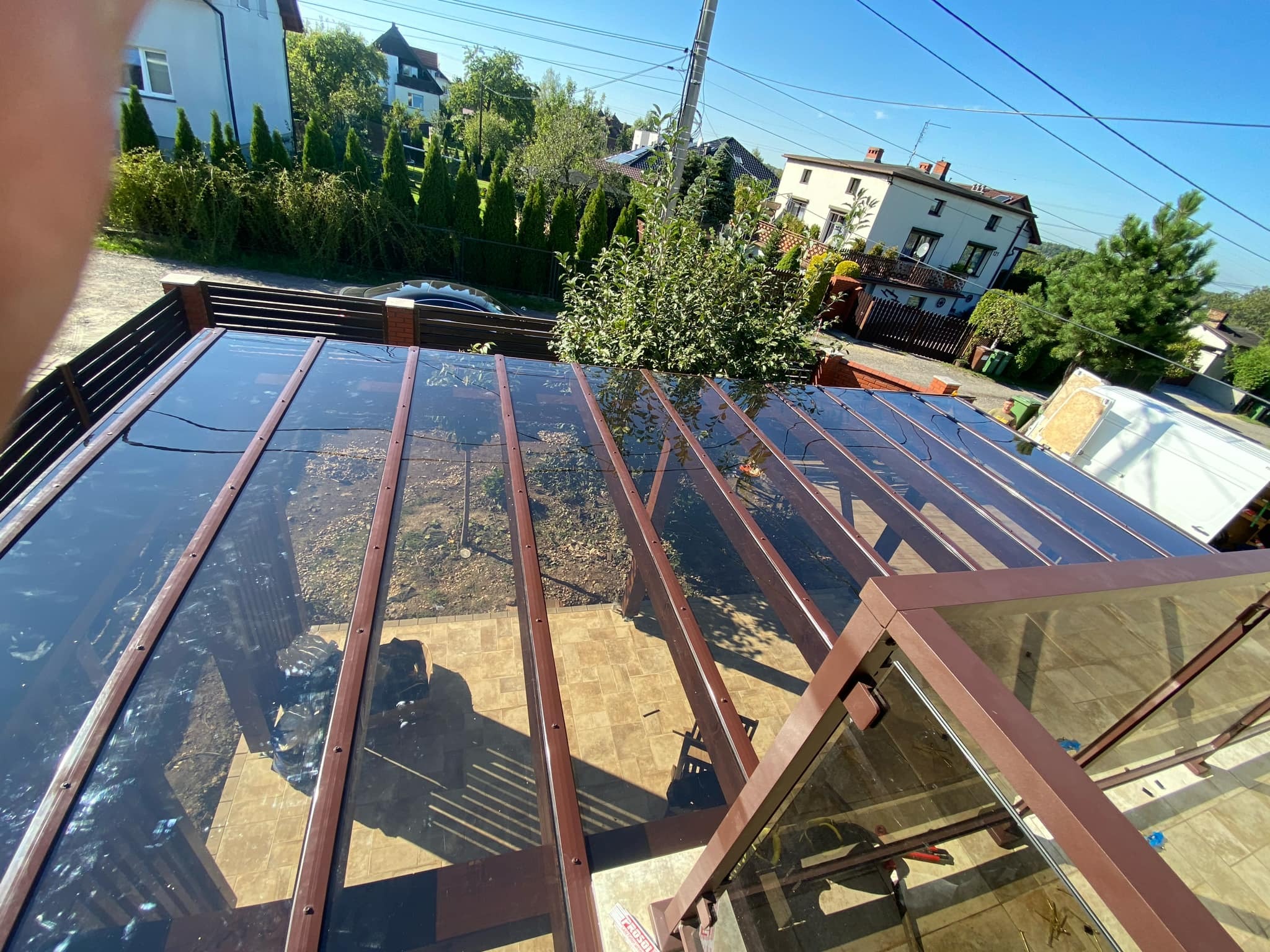 Szklana konstrukcja z brązowymi elementami nośnymi, prawdopodobnie zadaszenie tarasu, widziana z góry, z elementami otoczenia w tle.