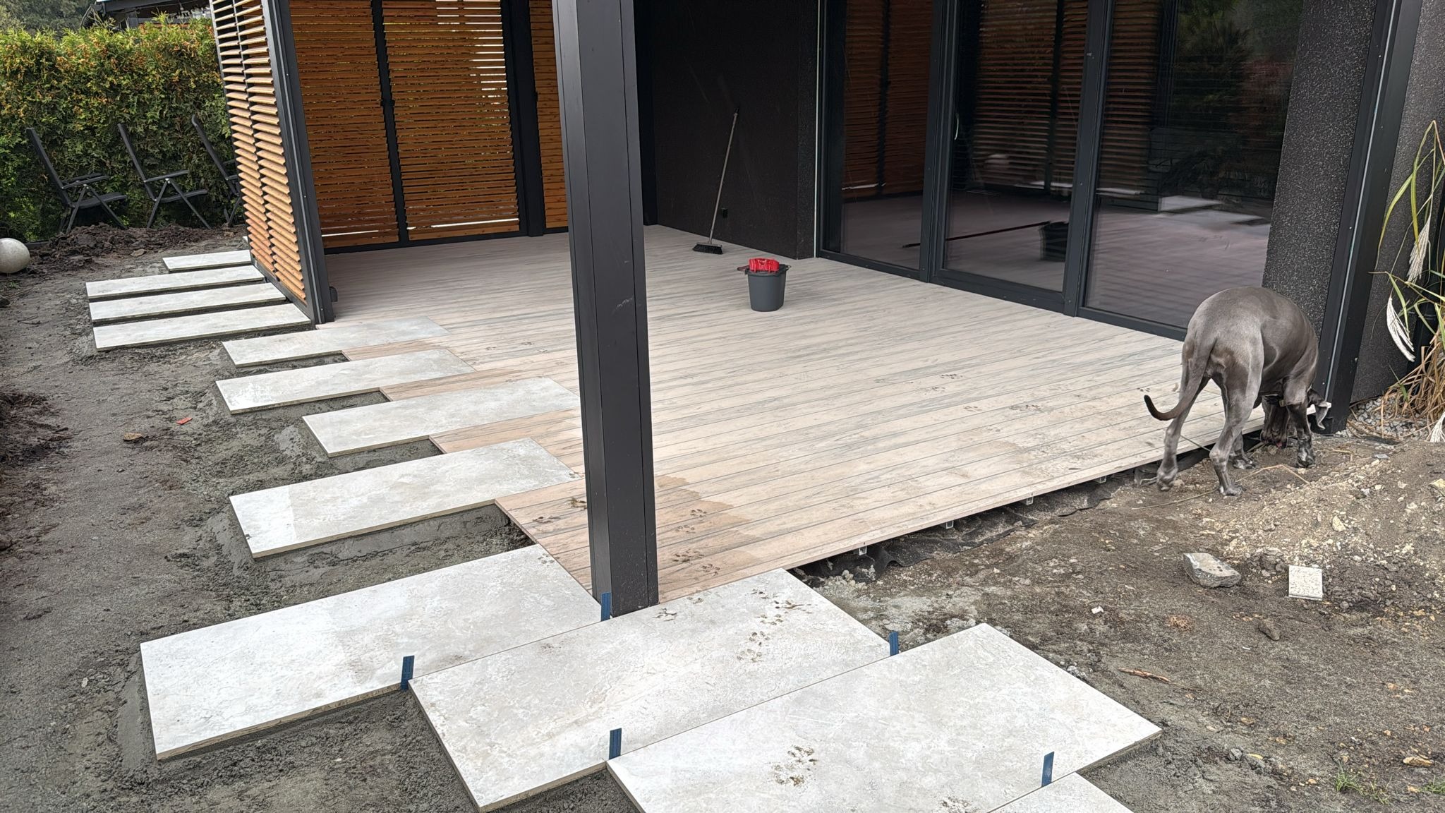 Montaż tarasu: deski i kamienne płyty przeplatane, pies Great Dane przygląda się pracy. Widoczny fragment budynku z drewnianymi żaluzjami i czarnymi ramami okien.