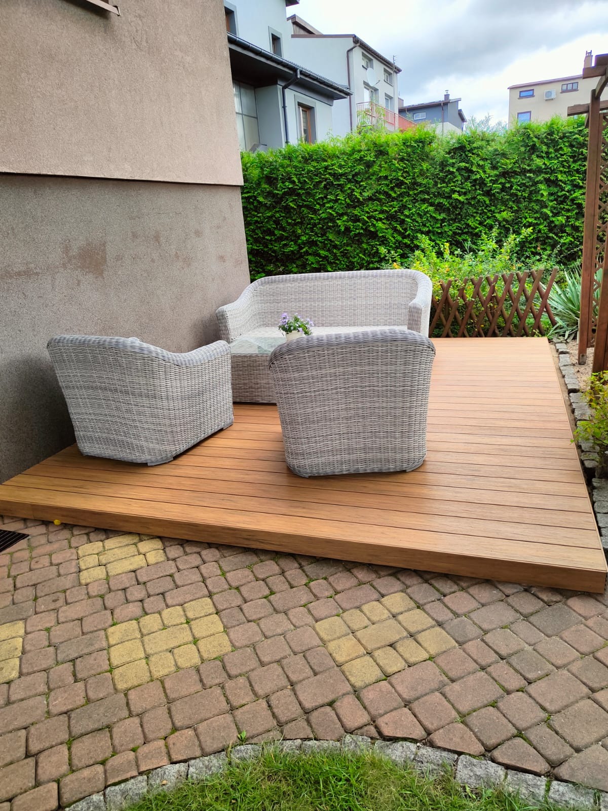 Nowoczesny taras z desek kompozytowych, otoczony kostką brukową. Wygodne meble ogrodowe z rattanu, zielony żywopłot w tle. Słoneczny dzień.