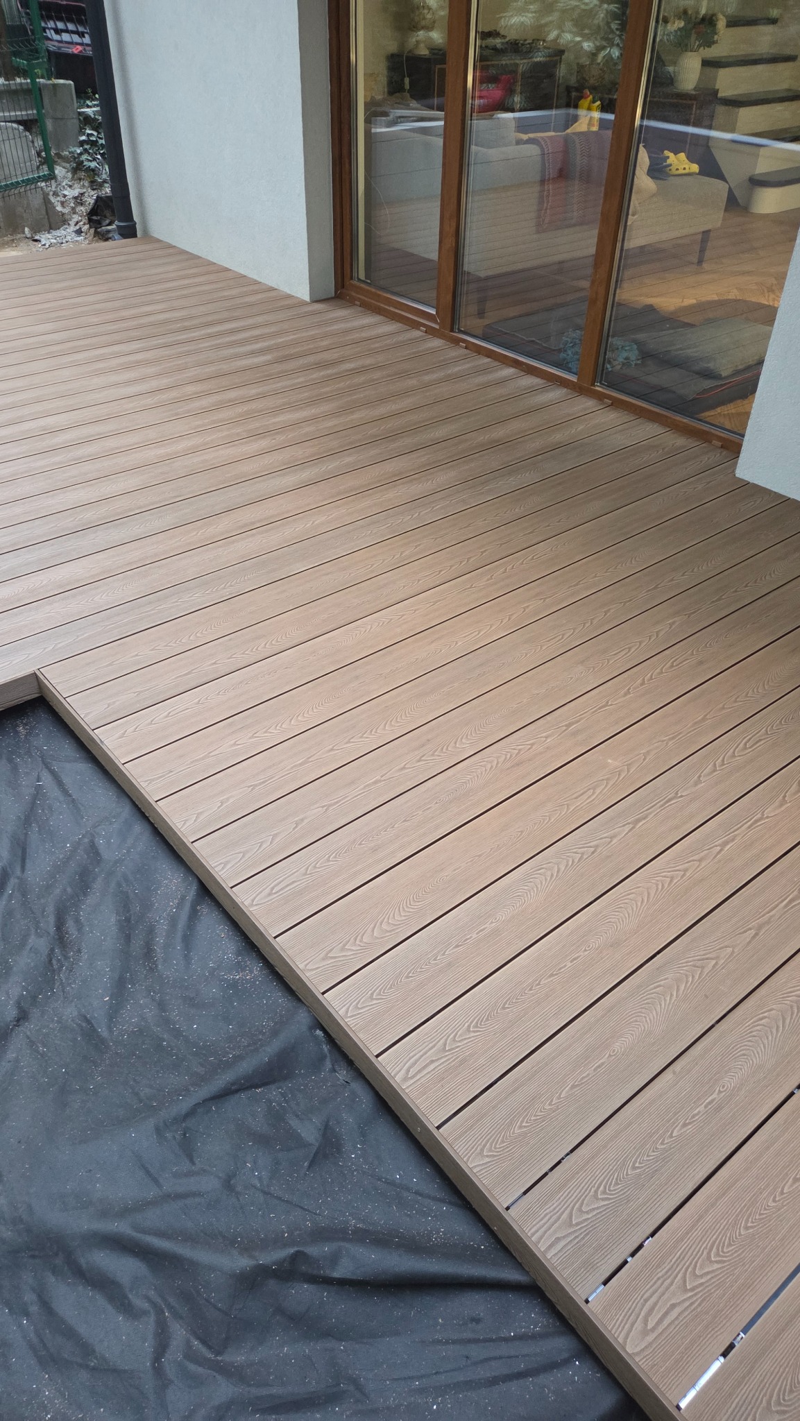 Jasny taras kompozytowy z widoczną konstrukcją pod spodem, częściowo zakrytą czarną folią, przylegający do budynku z dużym oknem.