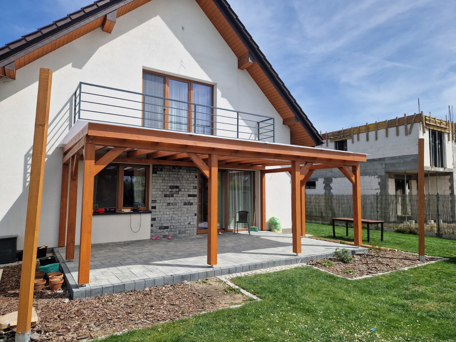 Drewniana pergola z jasnego drewna przylegająca do domu z balkonem, na tle zielonego trawnika i budowy. Widoczne donice i narzędzia.