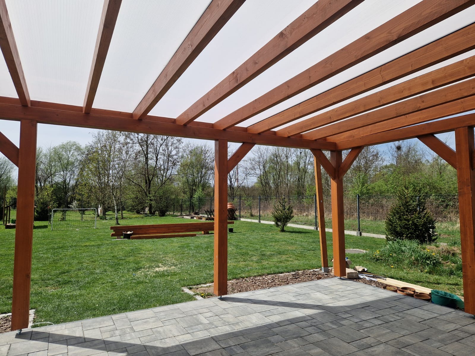 Drewniana pergola z transparentnym zadaszeniem, widok z poziomu tarasu z kostki brukowej na zielony ogród z bramką piłkarską w tle.