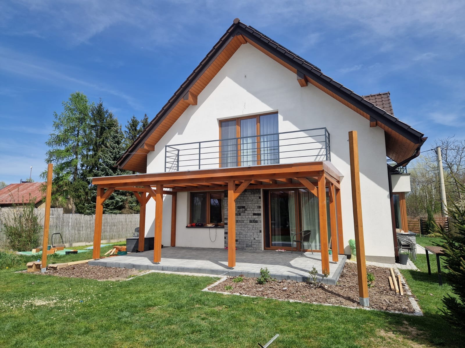 Dom z drewnianą pergolą w trakcie budowy, widoczne słupy i zadaszenie. Częściowo wyłożony taras z kostki brukowej. Widok na ogród i błękitne niebo.