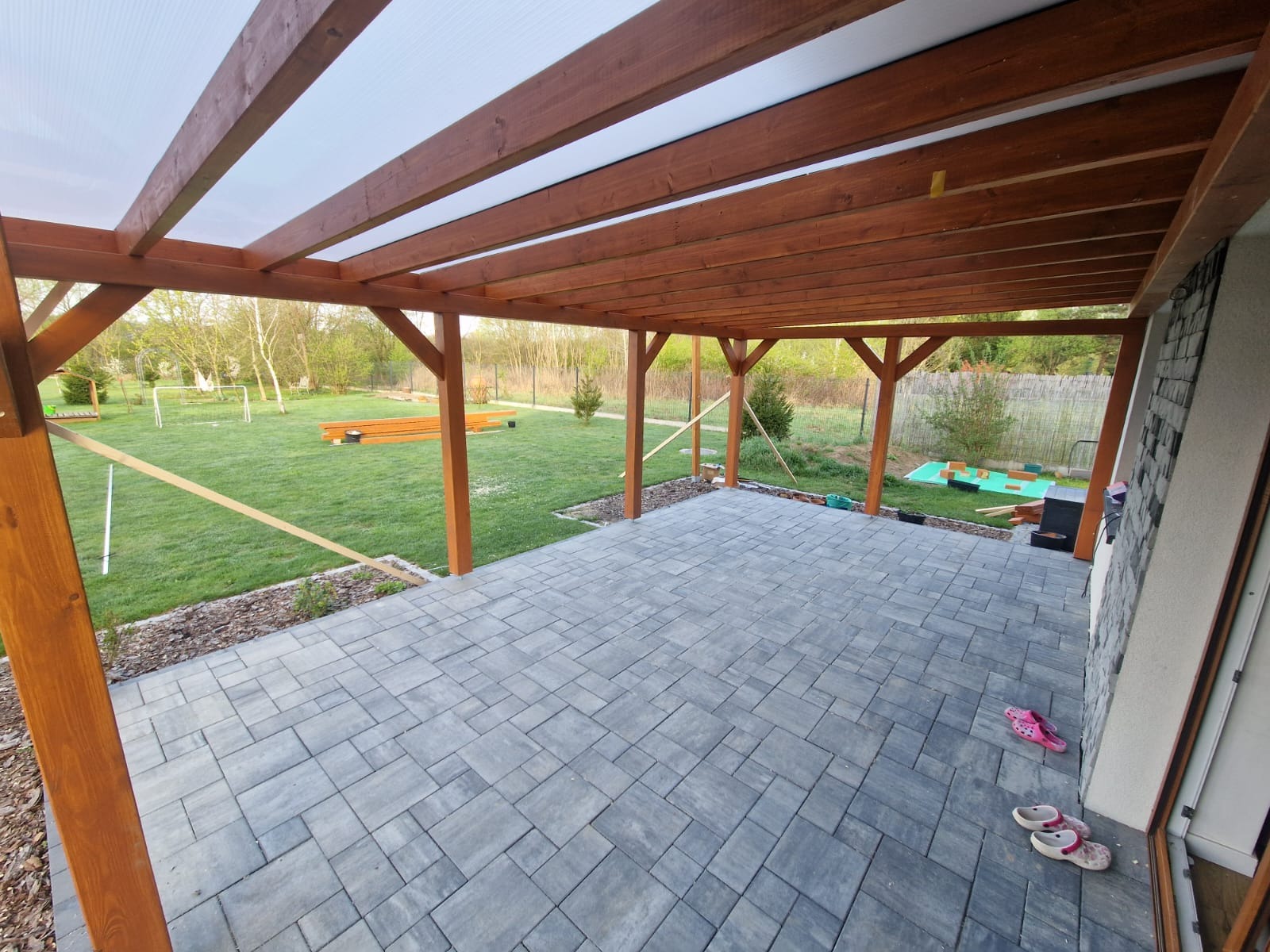 Zadaszony taras z szarej kostki brukowej i drewnianą konstrukcją. W tle zielony trawnik i drzewa. Na tarasie leżą klapki.
