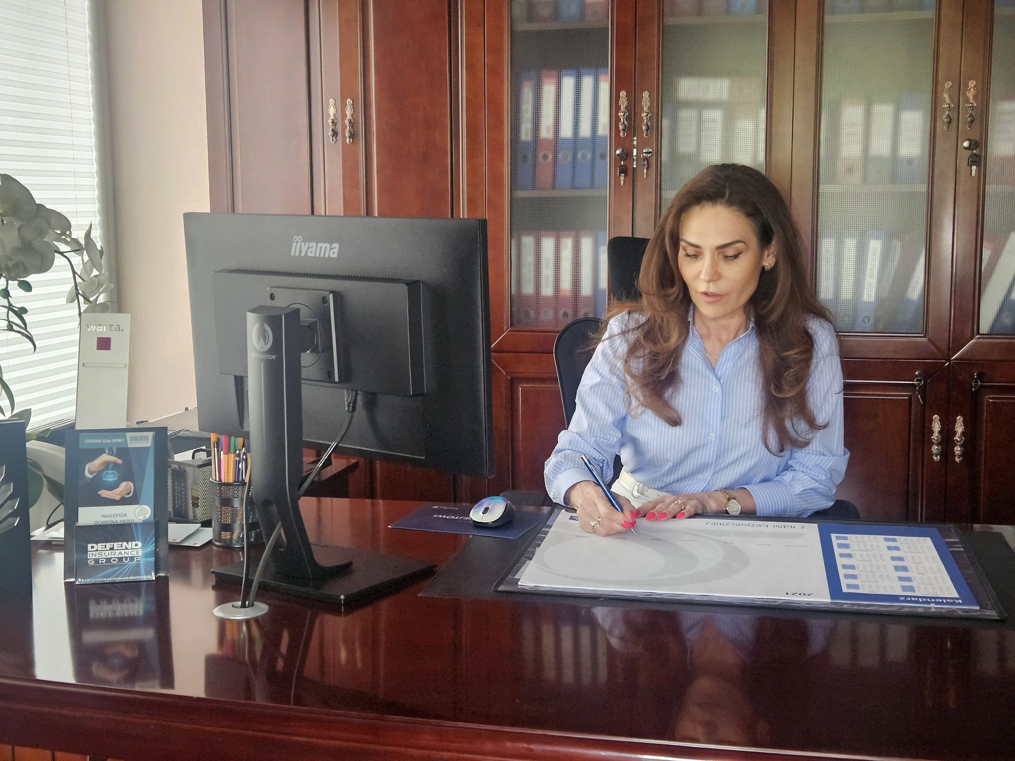 Kobieta w biurze, podpisuje dokument na biurku z kalendarzem, widoczny monitor i broszura Defend Insurance Group
