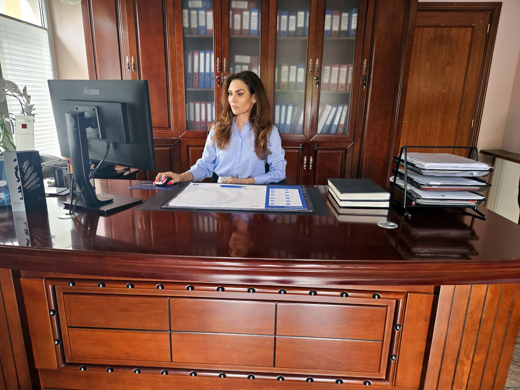 Kobieta w błękitnej koszuli siedzi za dużym, mahoniowym biurkiem, używając komputera w eleganckim gabinecie z regałami pełnymi segregatorów.