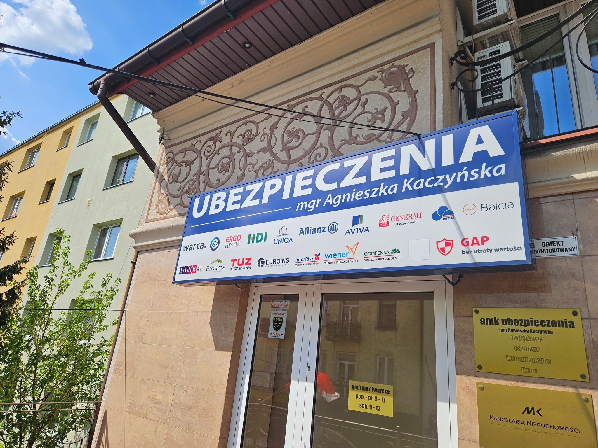 Szyld biura ubezpieczeniowego z nazwiskiem mgr Agnieszki Kaczyńskiej i logo różnych towarzystw ubezpieczeniowych, umieszczony nad wejściem do budynku z dekoracyjnym ornamentem.