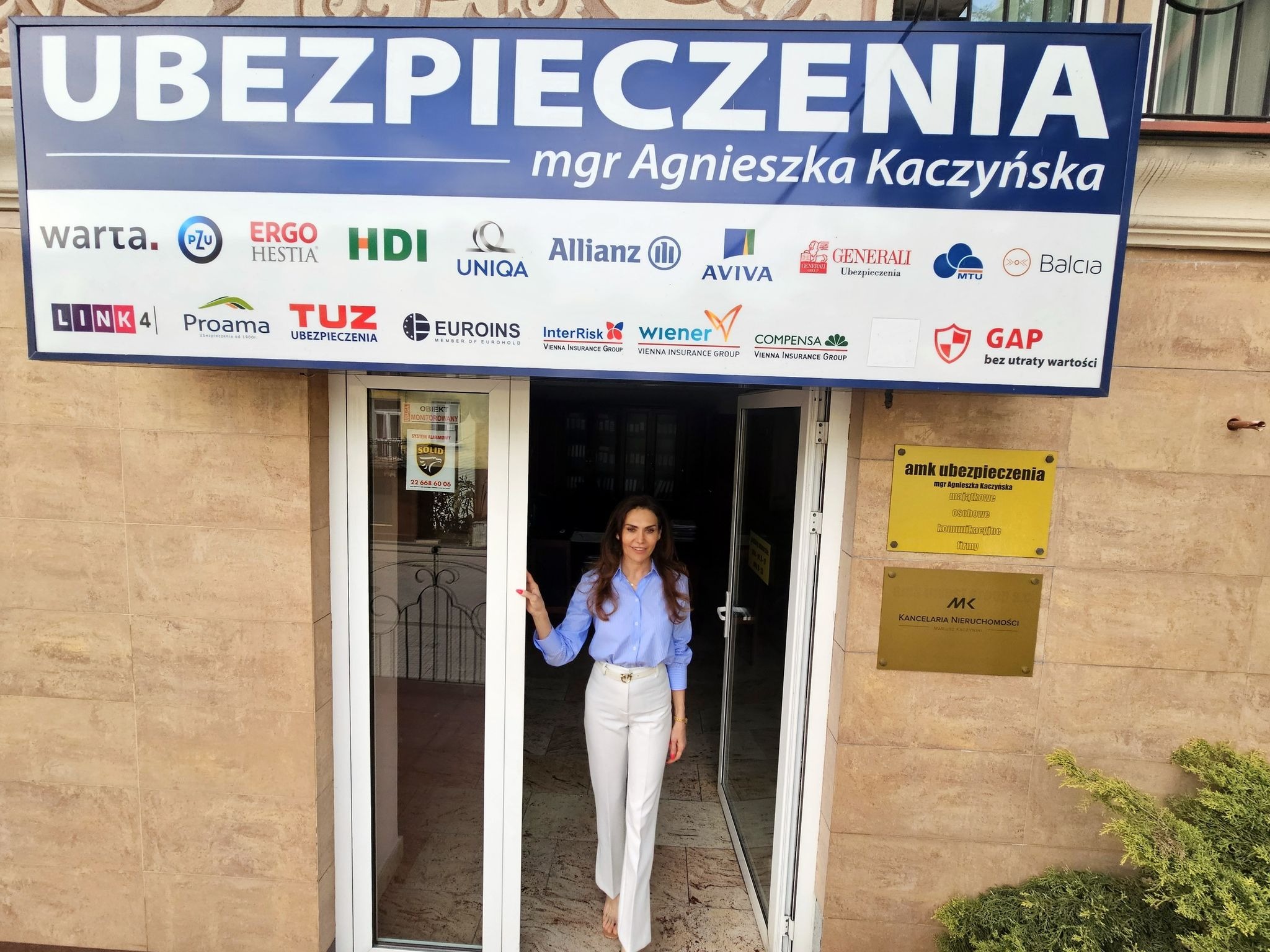 Kobieta w białych spodniach i błękitnej koszuli wychodzi z biura ubezpieczeń mgr Agnieszki Kaczyńskiej, nad wejściem szyld z logo wielu firm ubezpieczeniowych.