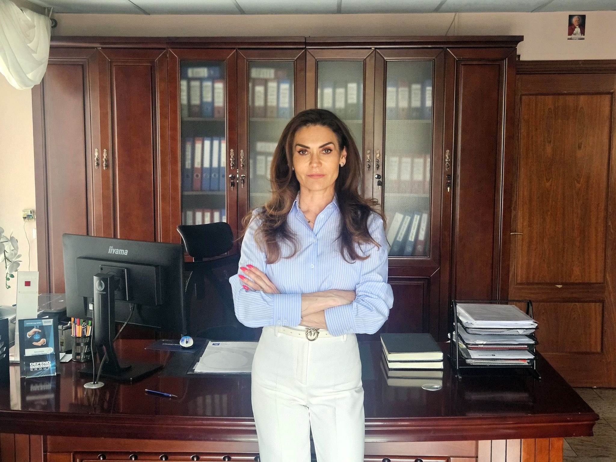 Kobieta w błękitnej koszuli i białych spodniach stoi z założonymi rękami na tle biura z ciemnymi meblami i segregatorami w szafkach.