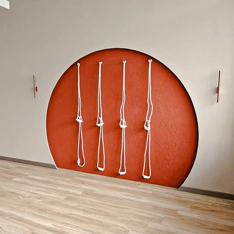 Ściana w neutralnym odcieniu z wkomponowanym pomarańczowym półokręgiem, na którym zamocowano cztery białe liny z węzłami, podłoga wyłożona panelami imitującymi jasne drewno, oświetlenie ścienne...