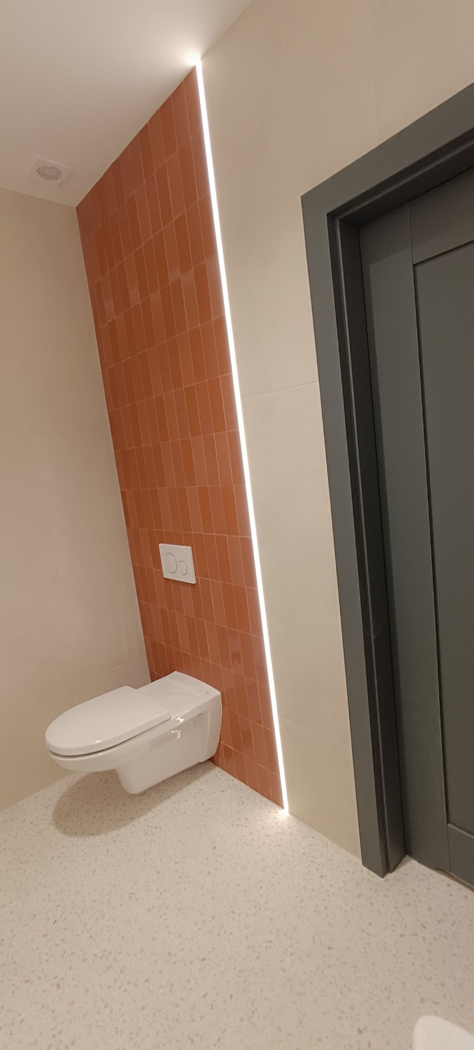 Nowoczesna toaleta z podświetlaną ścianą z cegiełek, wisząca miska WC i szara ościeżnica drzwiowa na tle podłogi z lastryko.