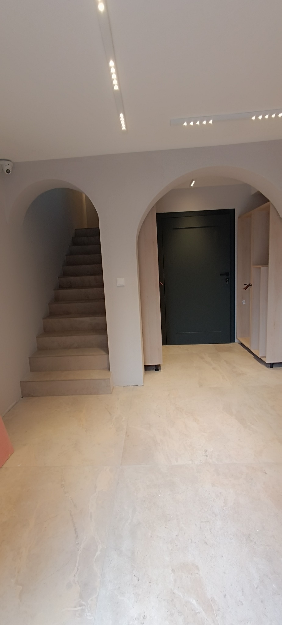 Jasne wnętrze z marmurową podłogą, łukowym wejściem do klatki schodowej i ciemnozielonymi drzwiami, oświetlone listwami LED.