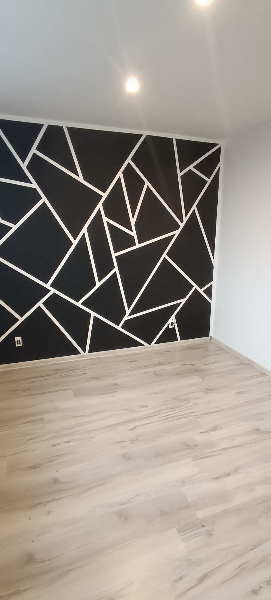 Czarna ściana z geometrycznym wzorem z białych linii w pustym pokoju z panelami podłogowymi i gniazdkami elektrycznymi.