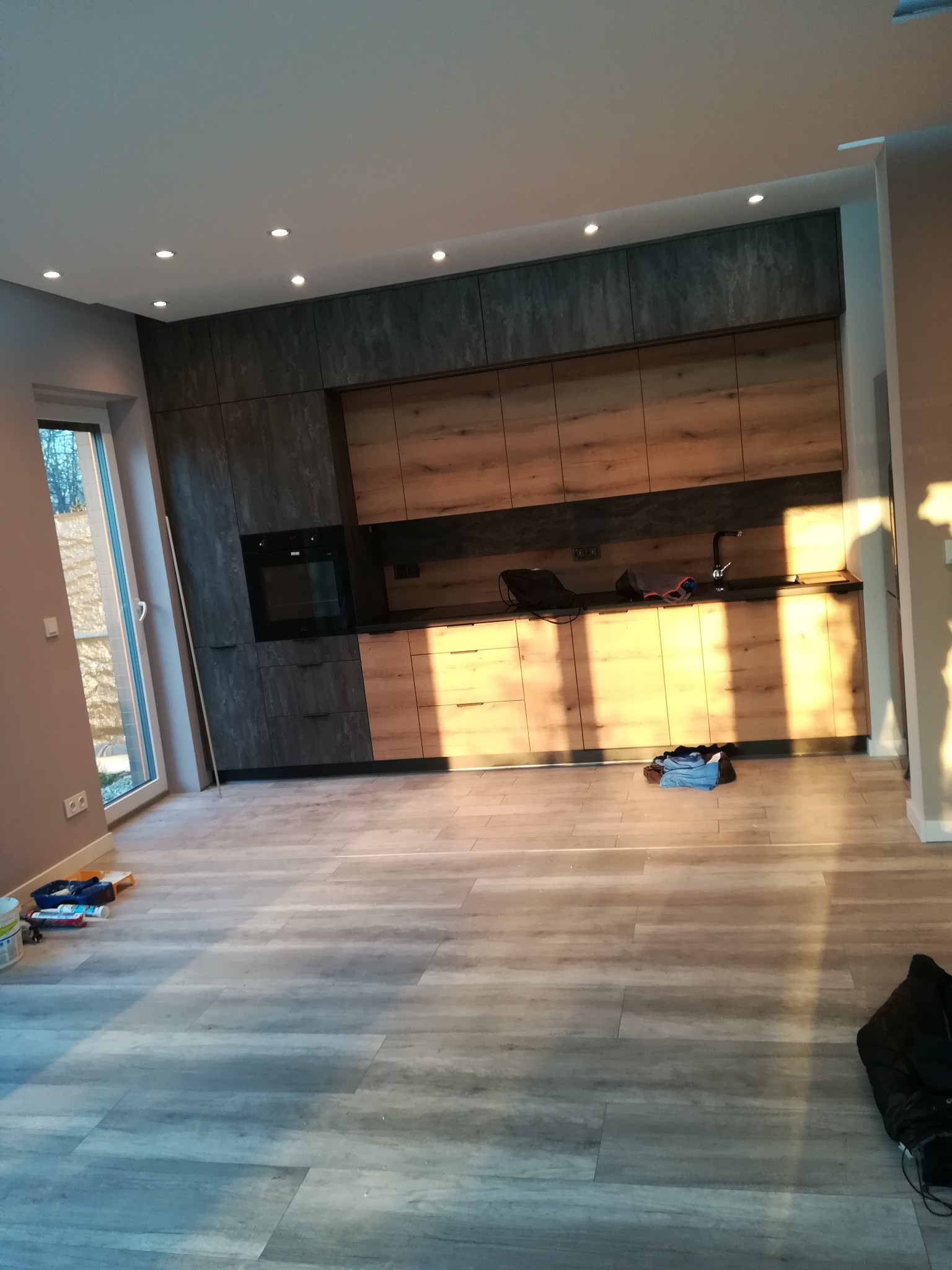 Nowoczesna kuchnia z drewnianymi i grafitowymi szafkami, jasną podłogą i punktowym oświetleniem sufitowym w trakcie wykańczania, widoczne narzędzia malarskie i odzież robocza.