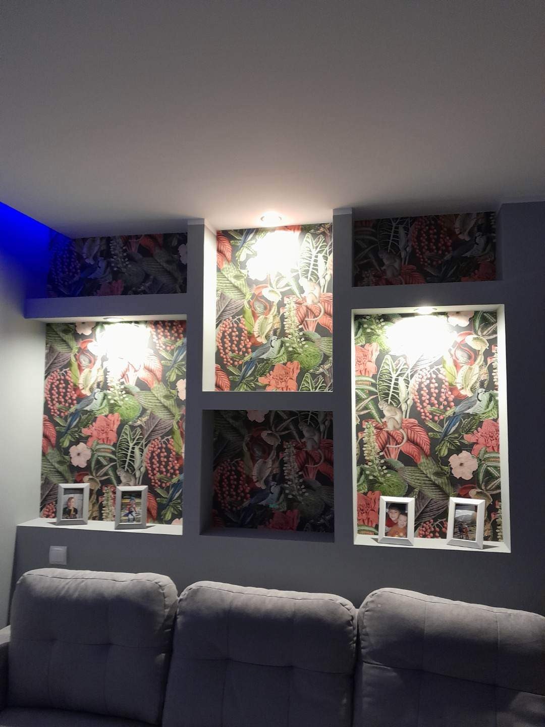 Wnęki w ścianie wykończone tapetą w tropikalne wzory, oświetlone punktowo, z dekoracyjnymi ramkami na półkach, widok z perspektywy sofy.