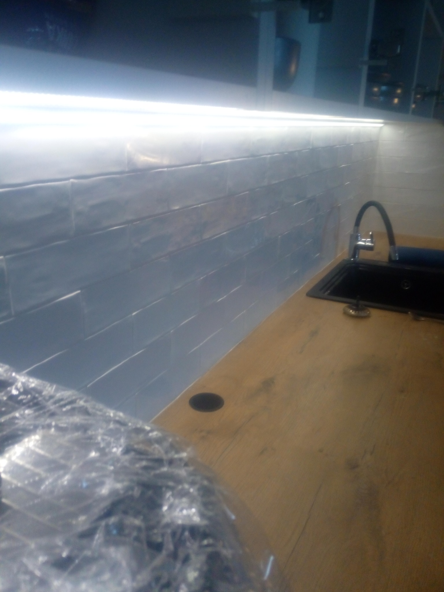 Kuchnia po remoncie: Błyszczące, szare płytki ułożone poziomo na ścianie nad blatem z drewnopodobnego laminatu, oświetlone ledowym paskiem, z czarnym zlewozmywakiem i chromowaną baterią.