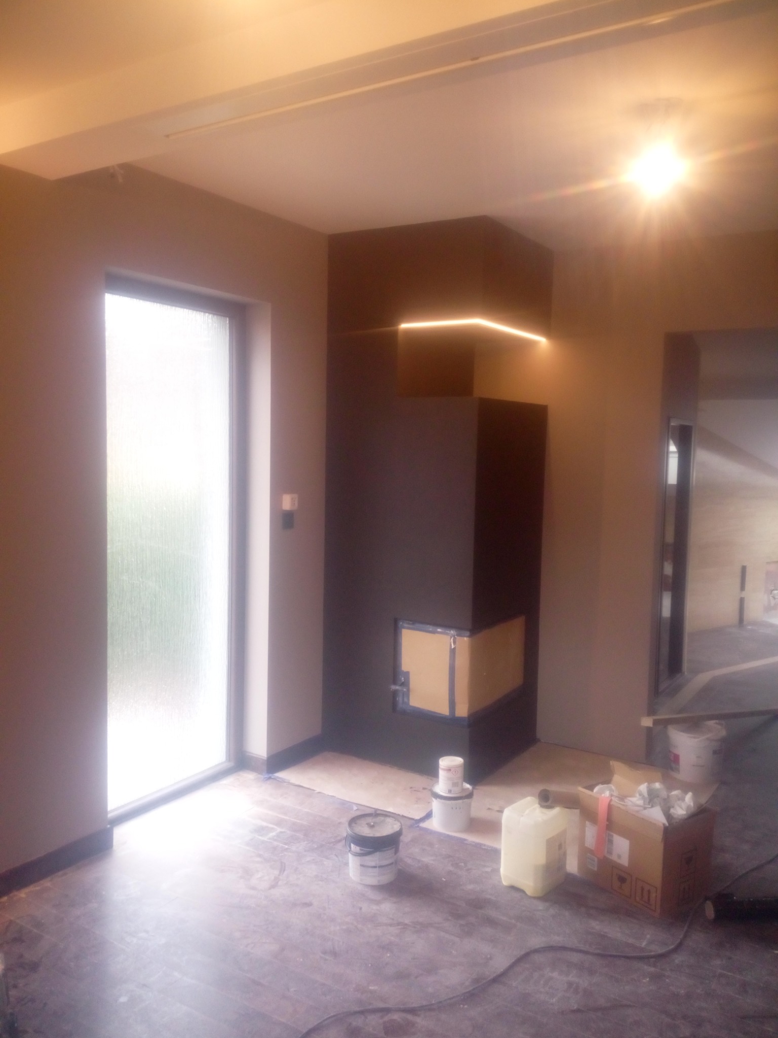 Wnętrze w trakcie remontu: pomalowane ściany w odcieniach szarości, nowoczesny kominek z oświetleniem LED, podłoga zabezpieczona folią, widoczne puszki z farbą i materiały budowlane.