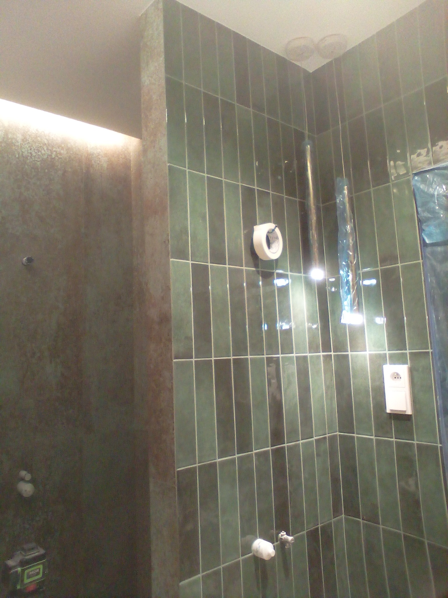 Wnętrze łazienki w trakcie prac: pionowe, ciemnozielone płytki ceramiczne na ścianach, fragment ściany z efektem betonu, widoczna taśma malarska, zabezpieczona folią rura i gniazdko elektryczne.