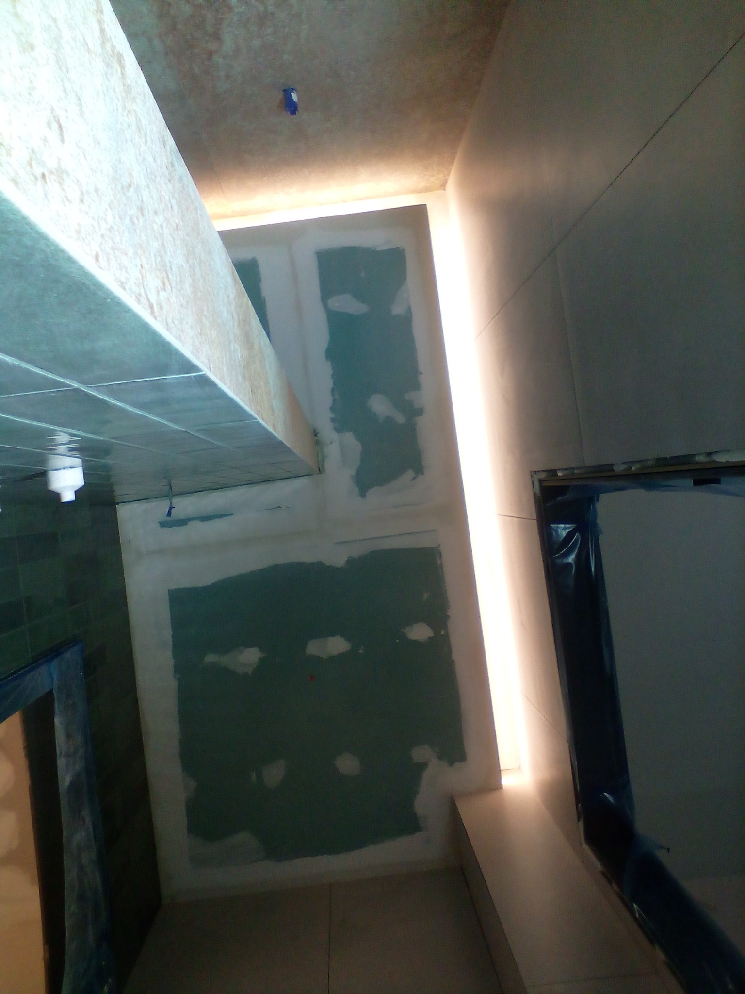 Ściana z płyt gipsowo-kartonowych w trakcie przygotowania do dalszych prac wykończeniowych, widoczne łączenia płyt pokryte masą szpachlową, oświetlenie LED wzdłuż ściany.