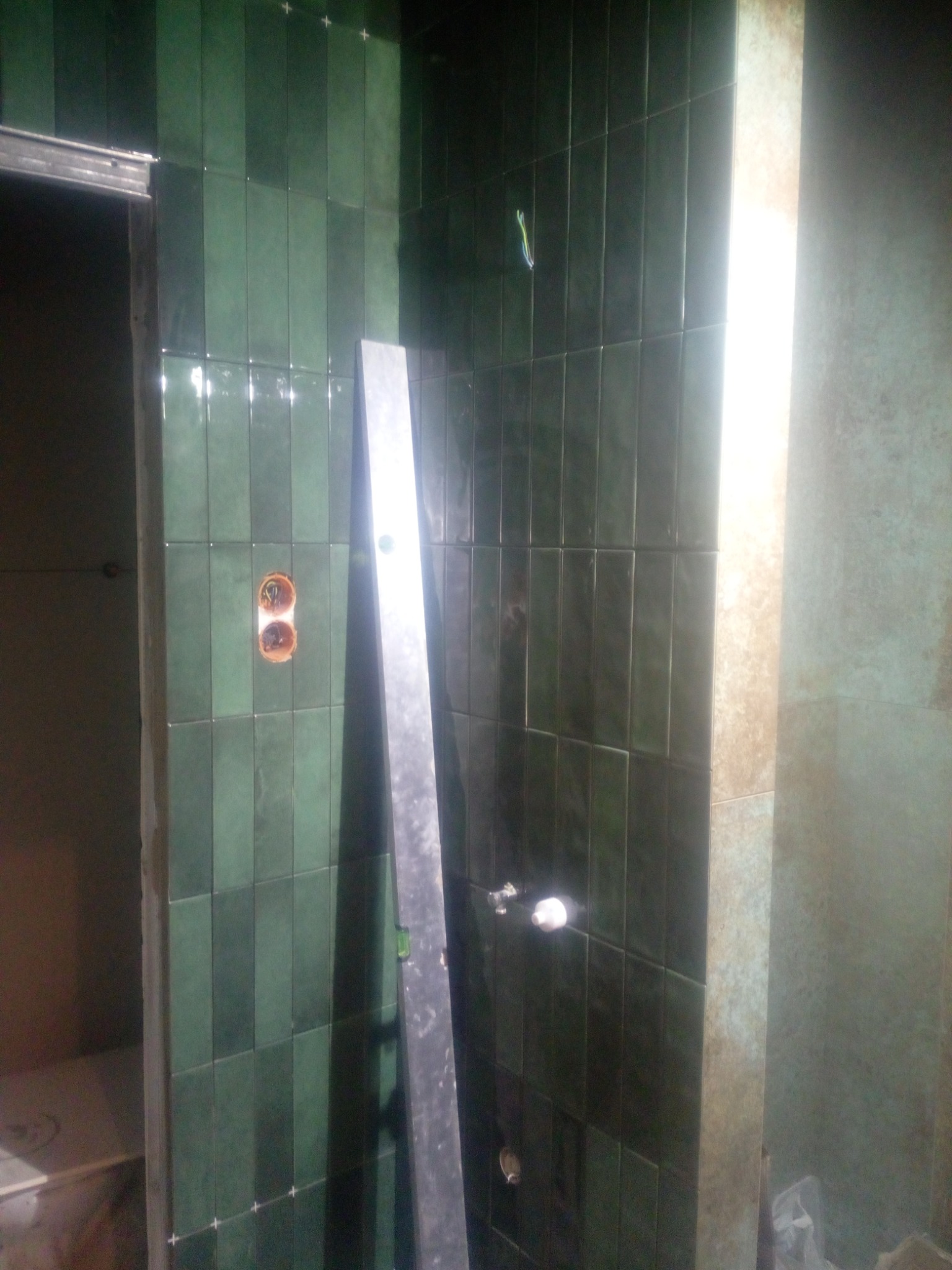 Montaż pionowych, błyszczących, zielonych płytek w łazience, widoczna poziomica przy ścianie, otwory na gniazdka i brakujące fragmenty fugi.