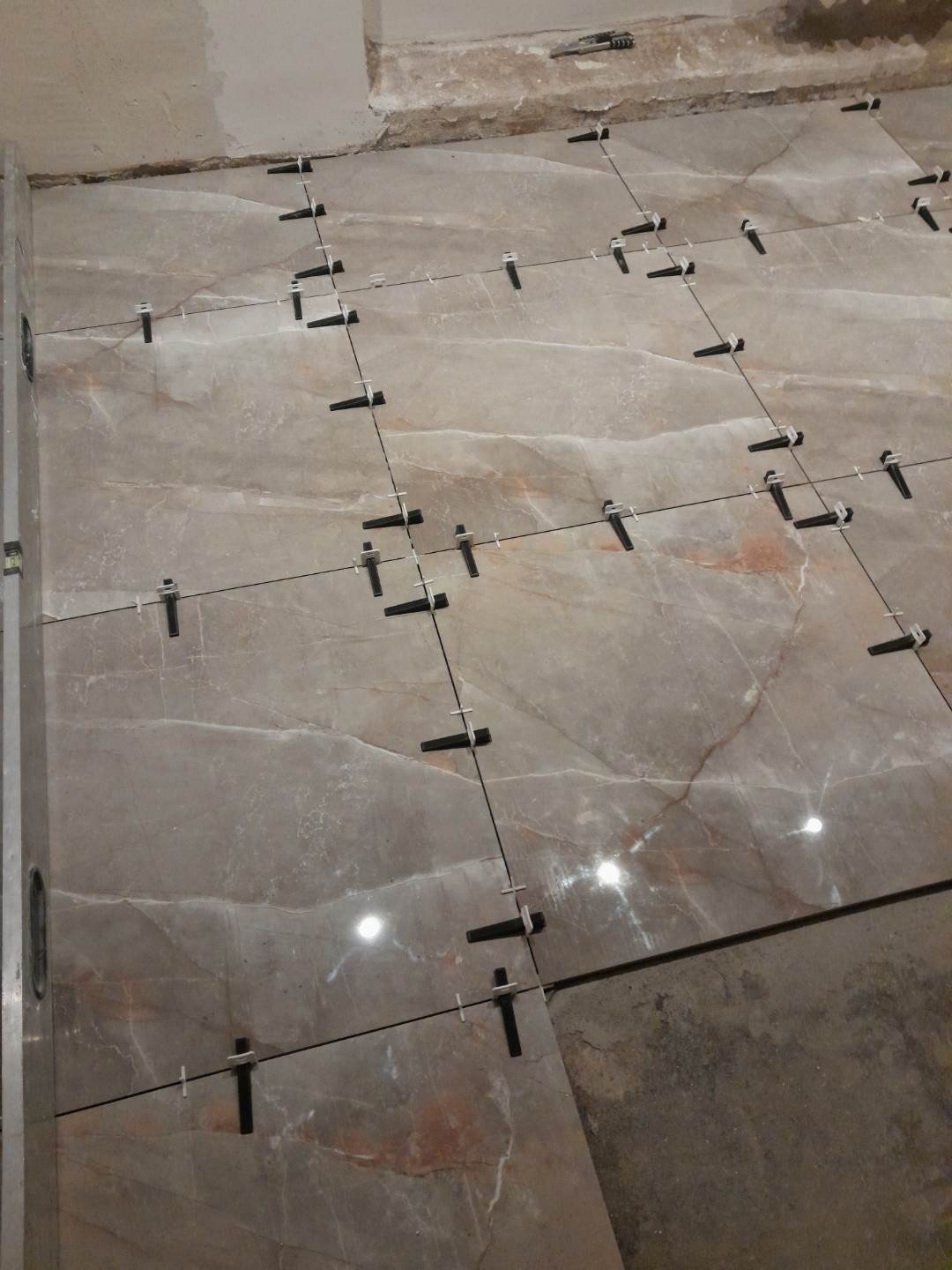 Układanie płytek podłogowych z użyciem systemu poziomowania; widoczne krzyżyki dystansowe i kliny, szare płytki z marmurkowym wzorem, surowy beton w tle.