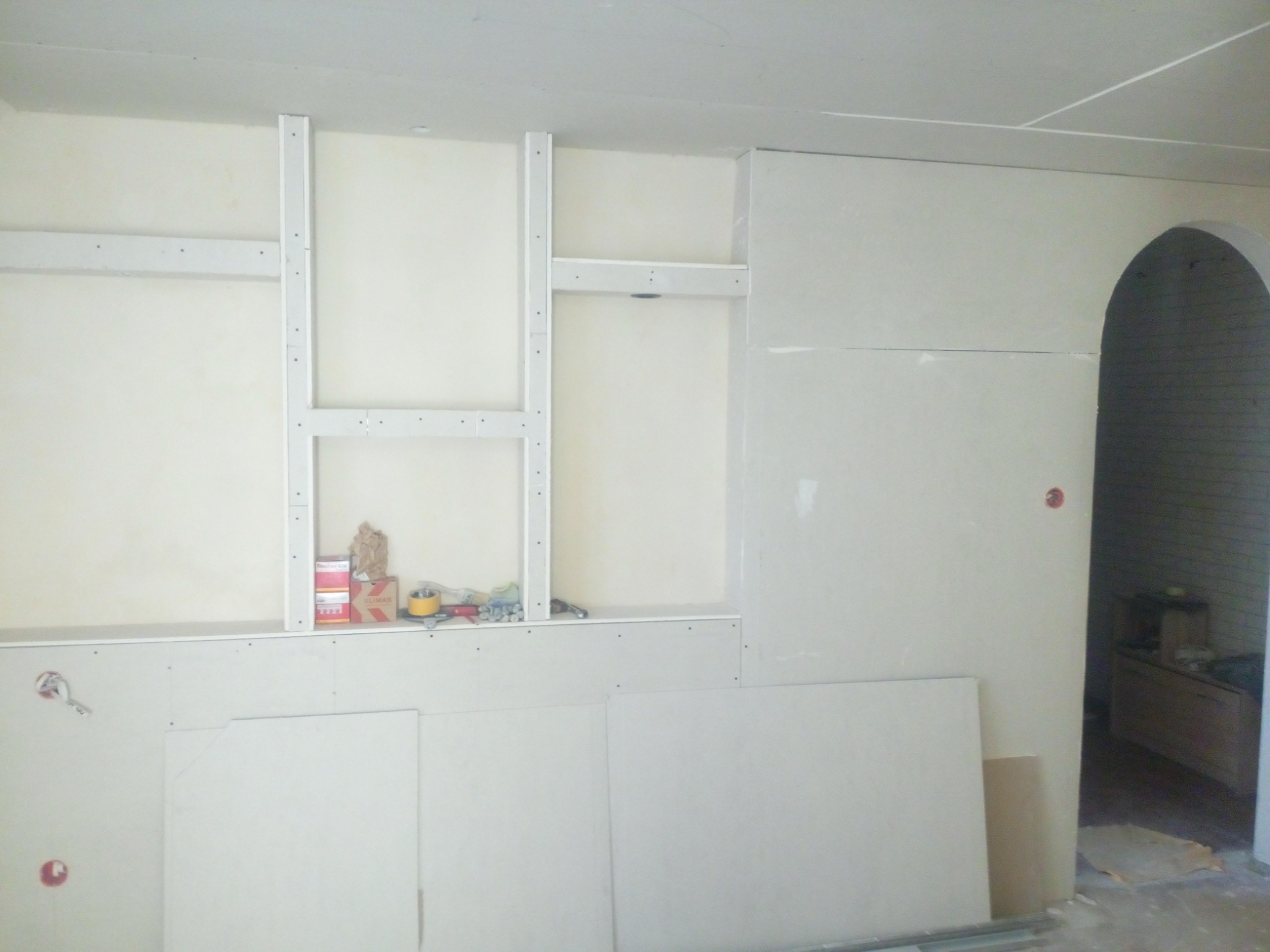 Montaż płyt gipsowo-kartonowych na ścianie z metalowym stelażem, widoczne narzędzia i materiały budowlane na półce, przygotowane otwory pod gniazdka elektryczne.