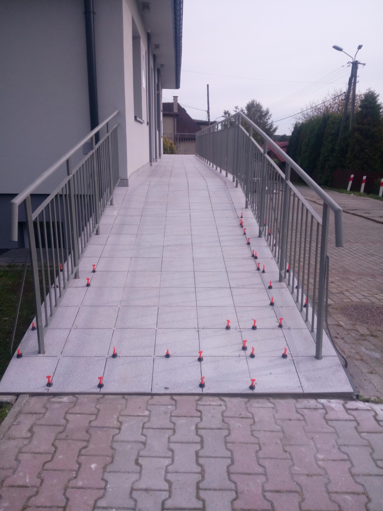 Rampa dla niepełnosprawnych w trakcie układania płytek, widoczne czerwone kliny poziomujące, metalowe barierki po bokach.