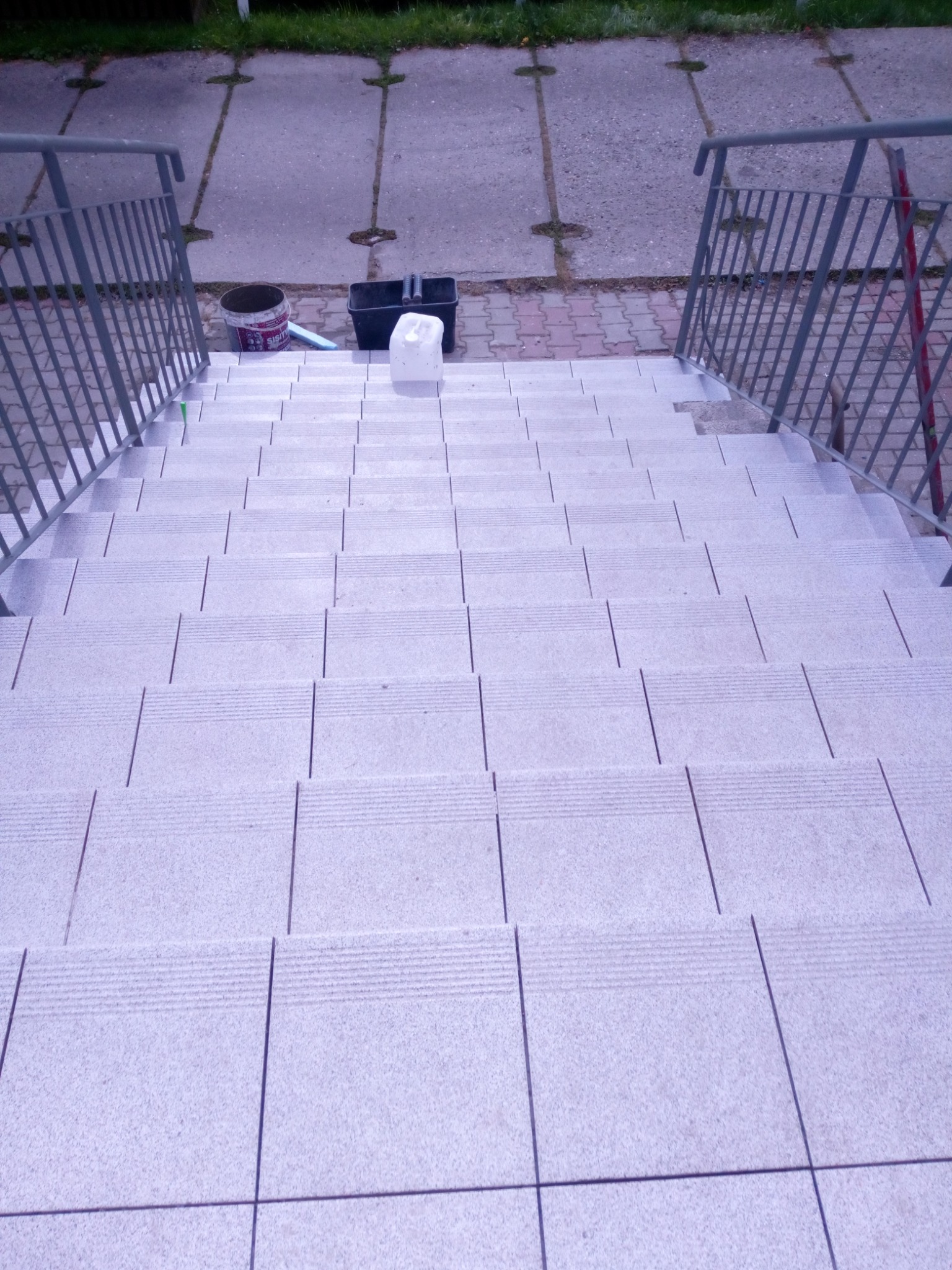 Widok z góry na schody wyłożone jasnymi płytkami, z metalową barierką po bokach, wiadro z farbą i narzędzia malarskie na górnym stopniu.