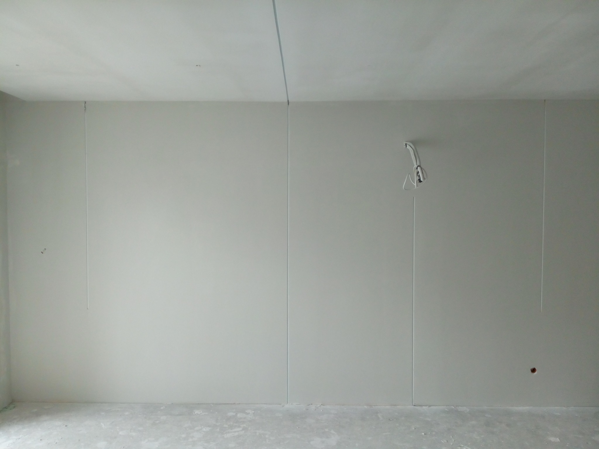 Przygotowana do malowania ściana z płyt gipsowo-kartonowych z widocznymi łączeniami, wystającymi przewodami elektrycznymi i surową podłogą, gotowa do dalszych prac wykończeniowych.