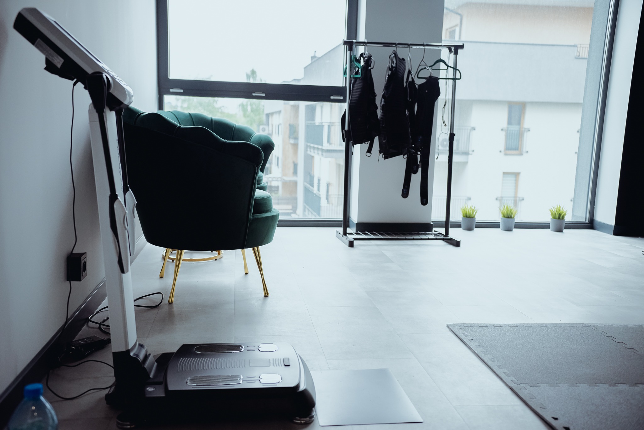 Wnętrze studia z maszyną do elektrostymulacji mięśni, zielonym fotelem na złotych nogach i wieszakiem z kombinezonami do EMS na tle okna z widokiem na miasto.