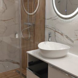 Artur Kowalewski - Nowoczesna łazienka z kabiną prysznicową ze szklaną ścianką, okrągłym lustrem z podświetleniem LED i białą umywalką nablatową na szafce z szufladami.