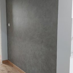 Artur Kowalewski - Ściana z efektem betonu w odcieniach szarości, drewniana podłoga, biała listwa przypodłogowa i fragment instalacji elektrycznej wystający z sufitu.