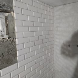 Artur Kowalewski - Ściana w trakcie wykańczania z białymi, błyszczącymi płytkami typu metro ułożonymi w cegiełkę, obok surowy beton i okno w trakcie montażu.