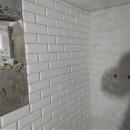 Ściana w trakcie wykańczania z białymi, błyszczącymi płytkami typu metro ułożonymi w cegiełkę, obok surowy beton i okno w trakcie montażu.