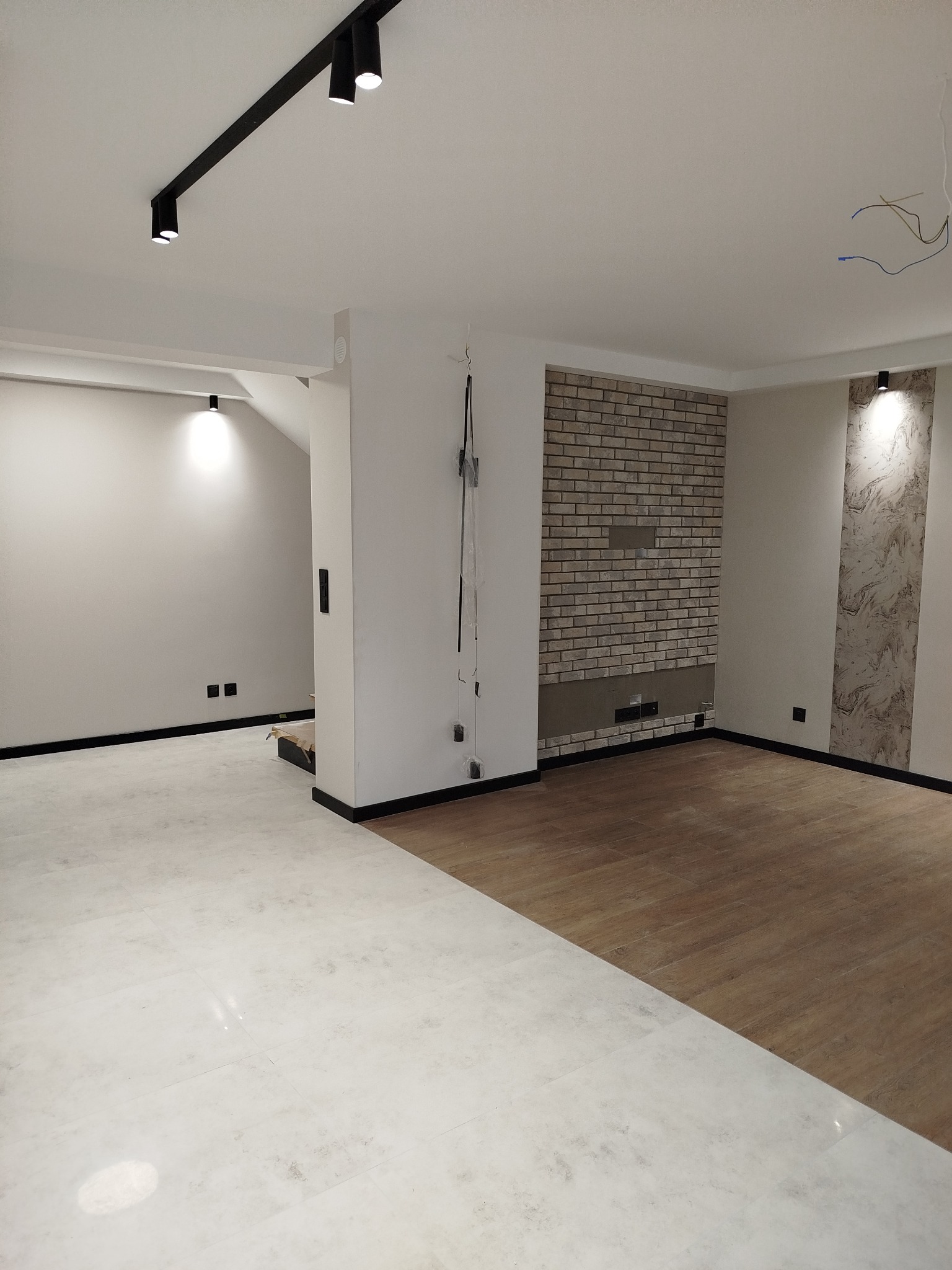 Wnętrze w trakcie prac wykończeniowych: połączenie podłogi z płytek imitujących marmur i paneli drewnopodobnych, ściana z dekoracyjnej cegły, sufit z szynoprzewodem oświetleniowym, widoczne kable...