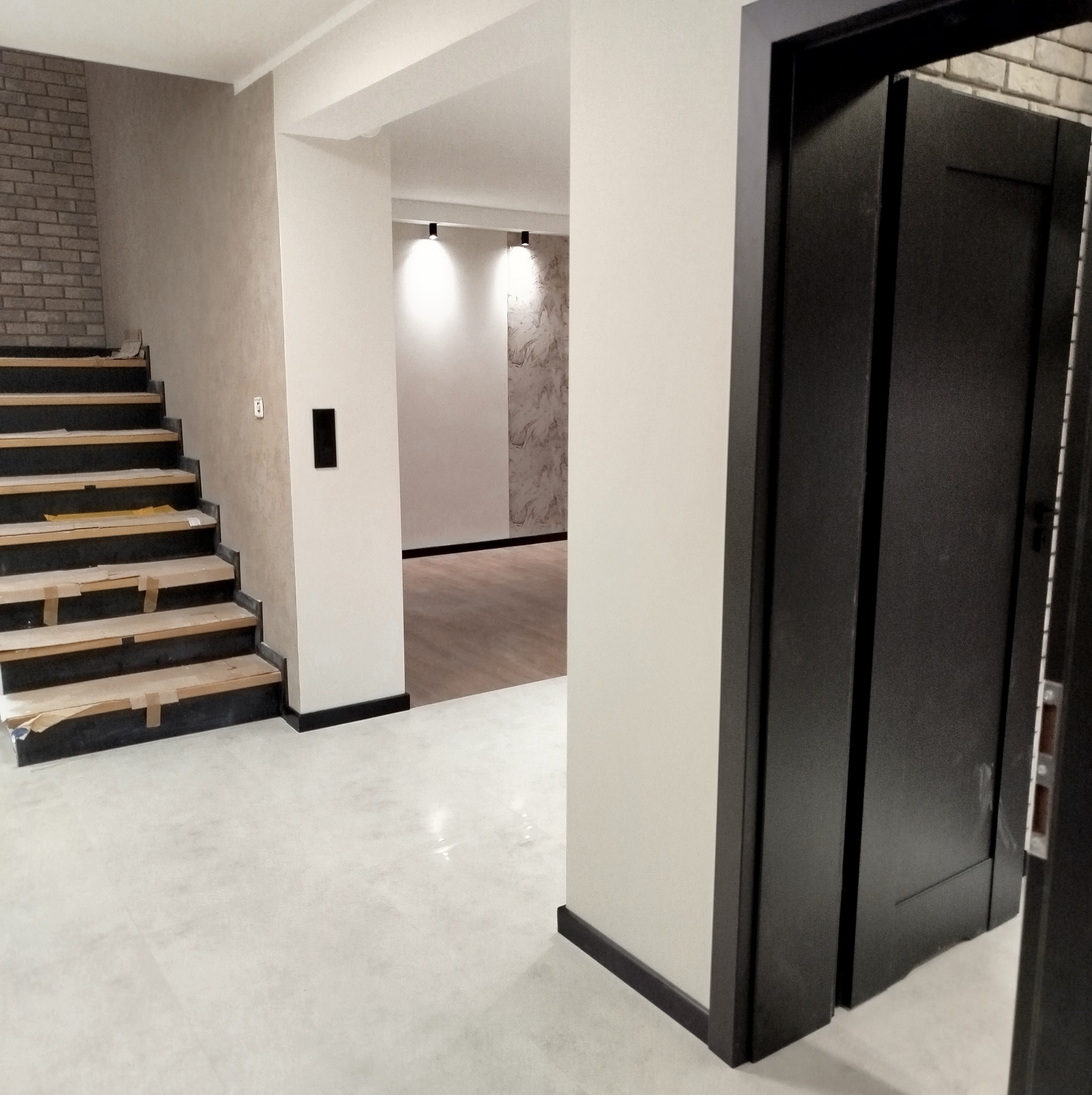 Wnętrze w trakcie wykańczania: schody z drewnianymi stopniami zabezpieczonymi folią, widoczny fragment ściany z cegły dekoracyjnej, podłoga wyłożona jasnymi płytkami, czarne listwy przypodłogowe...