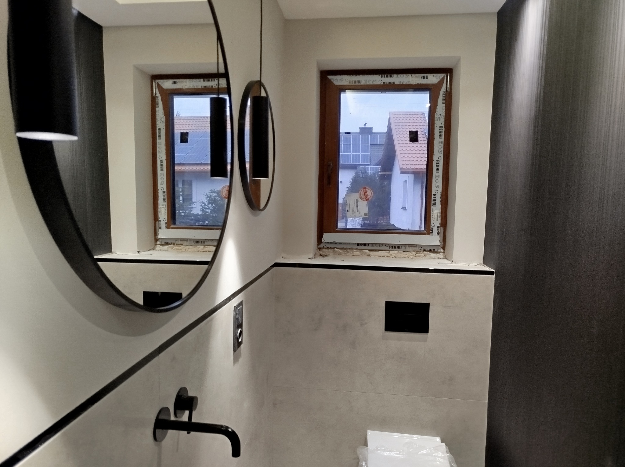 Nowoczesna łazienka w trakcie wykańczania, widoczne okna z widokiem na domy z panelami słonecznymi, czarna armatura, okrągłe lustro i lampy wiszące, płytki imitujące kamień z czarnymi akcentami.