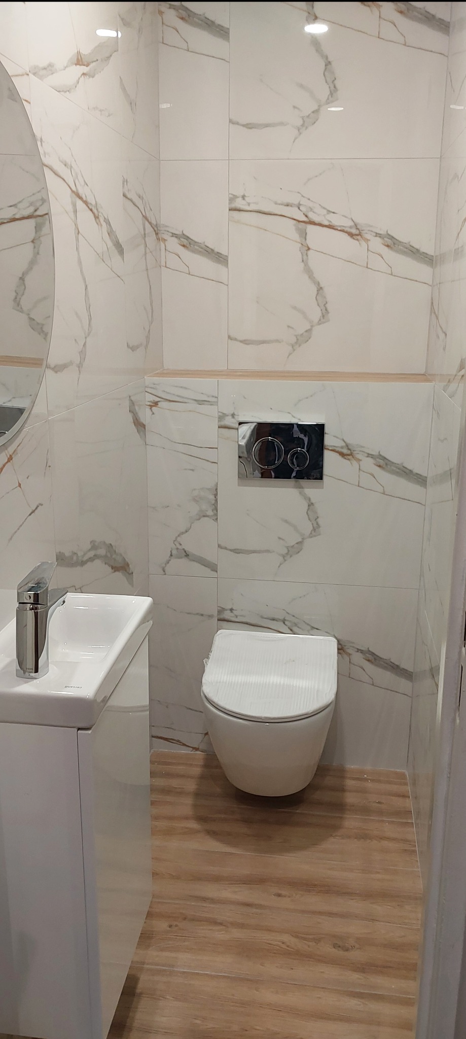 Nowoczesna łazienka z białymi płytkami marmurkowymi, podwieszaną toaletą, umywalką na szafce i drewnopodobną podłogą.