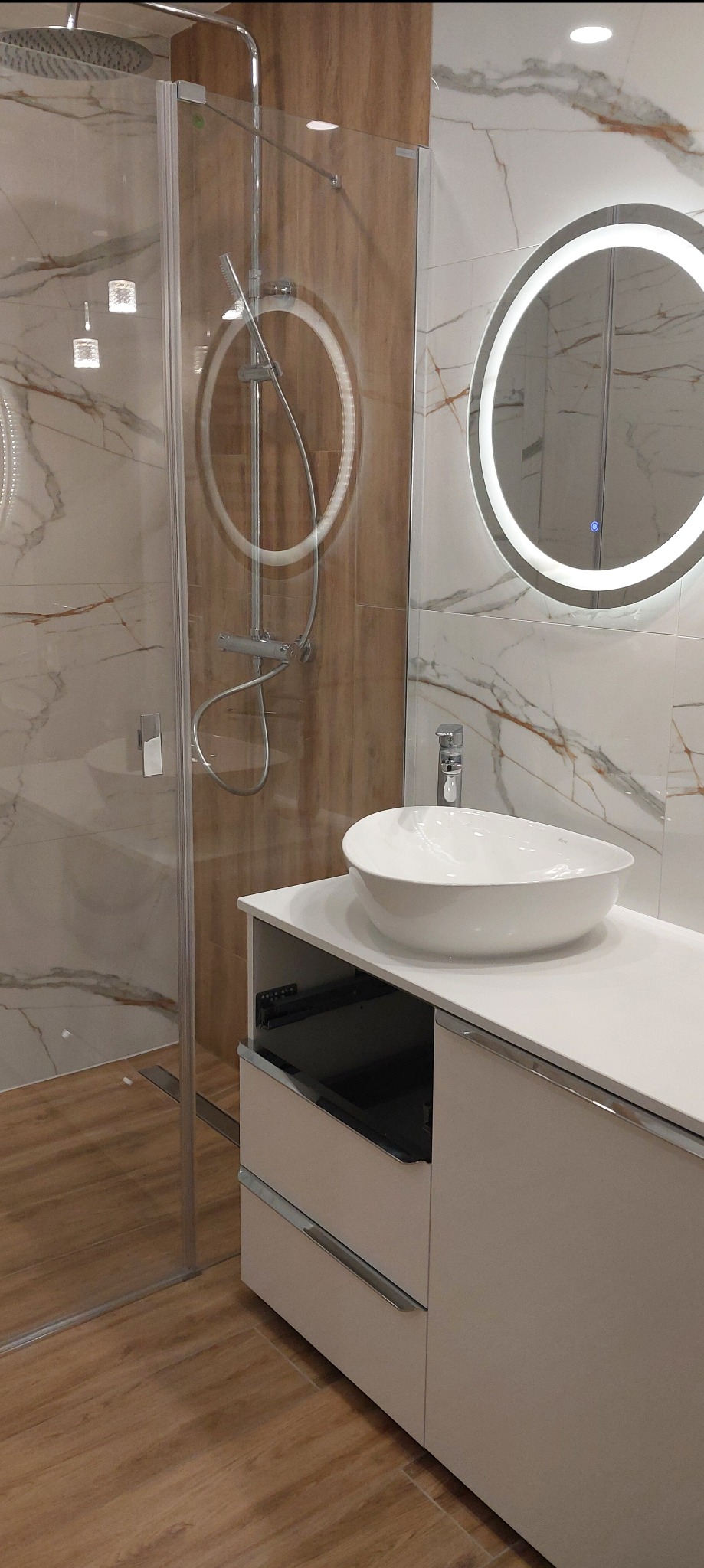 Nowoczesna łazienka z kabiną prysznicową ze szklaną ścianką, okrągłym lustrem z podświetleniem LED i białą umywalką nablatową na szafce z szufladami.