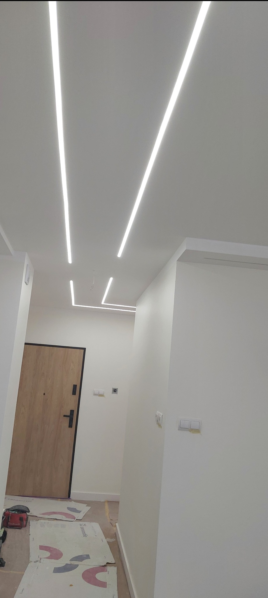 Nowoczesne oświetlenie sufitowe LED w korytarzu, z widocznymi listwami świetlnymi wpuszczonymi w sufit, zabezpieczone podłoże kartonami, drzwi wejściowe w tle.