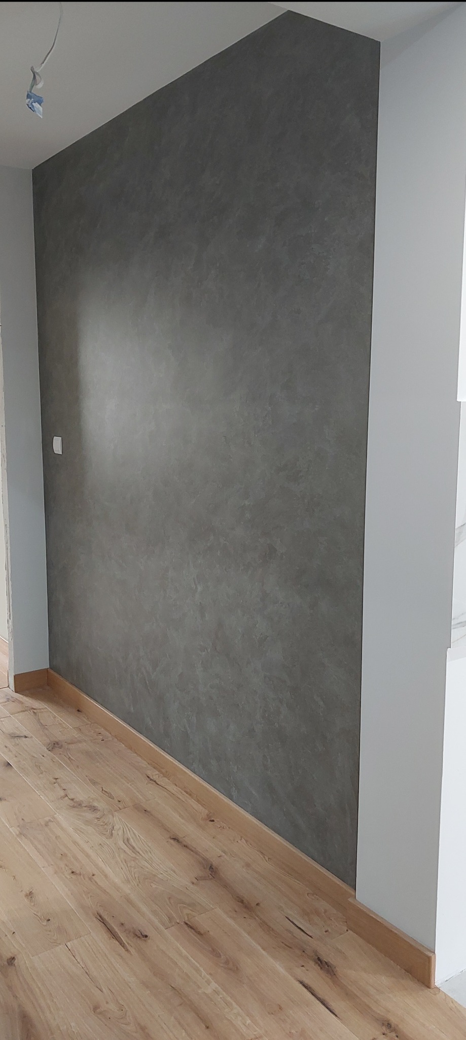 Ściana z efektem betonu w odcieniach szarości, drewniana podłoga, biała listwa przypodłogowa i fragment instalacji elektrycznej wystający z sufitu.