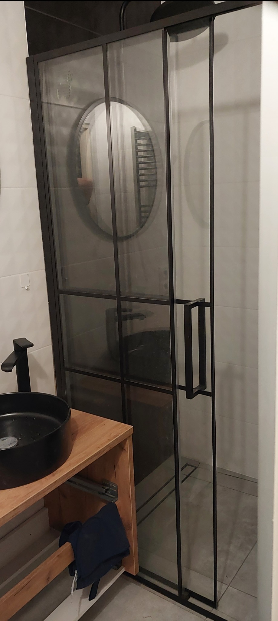 Nowoczesna łazienka z kabiną prysznicową typu walk-in z czarnymi profilami, okrągłym lustrem i czarną umywalką nablatową na drewnianej szafce.
