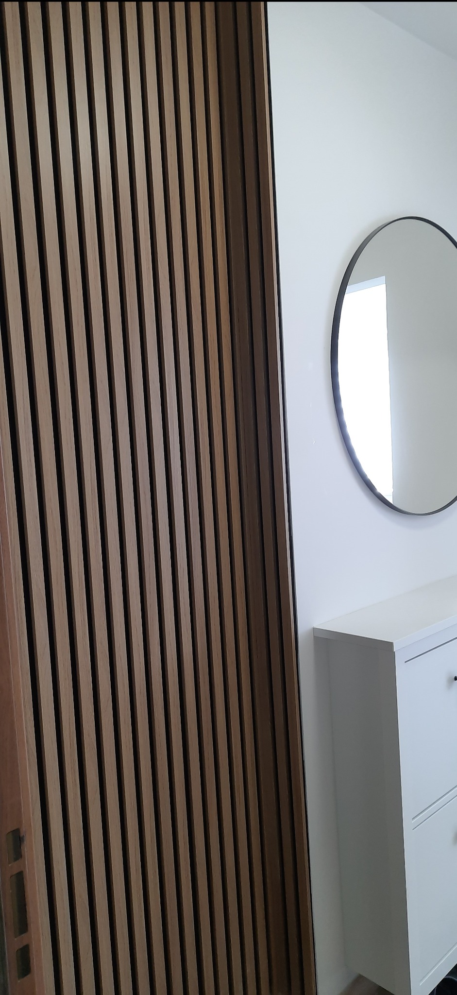 Ściana z drewnianych lameli w odcieniach brązu, obok biała komoda z okrągłym lustrem na ścianie.
