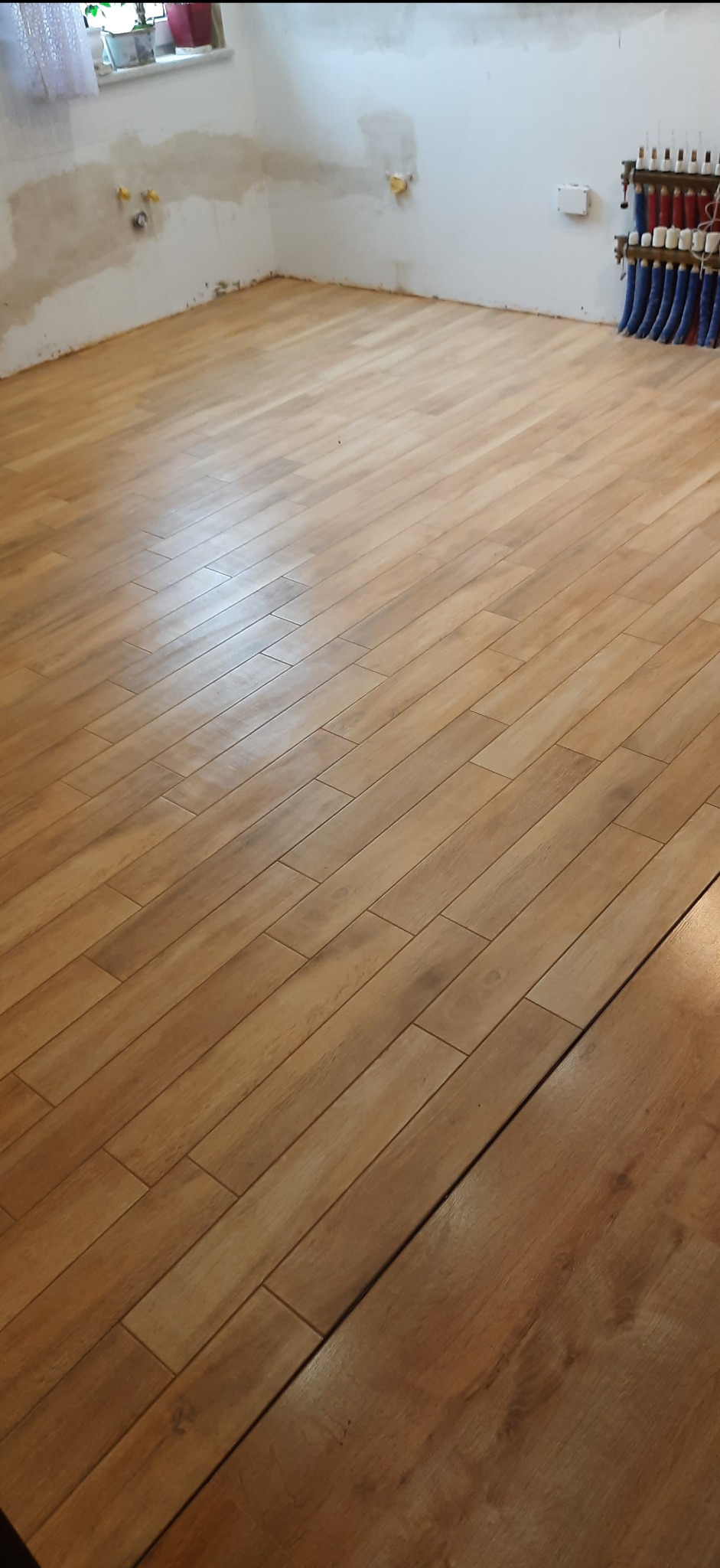 Ułożona podłoga z płytek imitujących drewno, widoczne niedokończone ściany z instalacją grzewczą i elektryczną w tle.