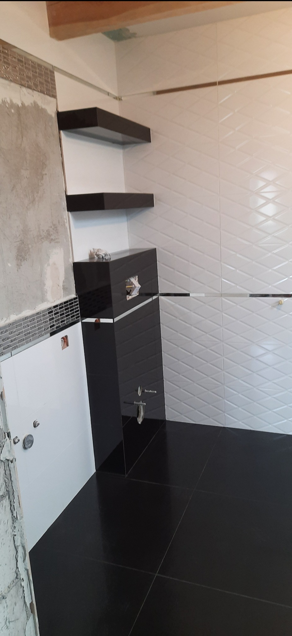 Częściowo wykończona łazienka z czarną podłogą i szafką oraz białymi ścianami z geometrycznym wzorem płytek, widoczne niedokończone fragmenty ściany z pozostałościami zaprawy.