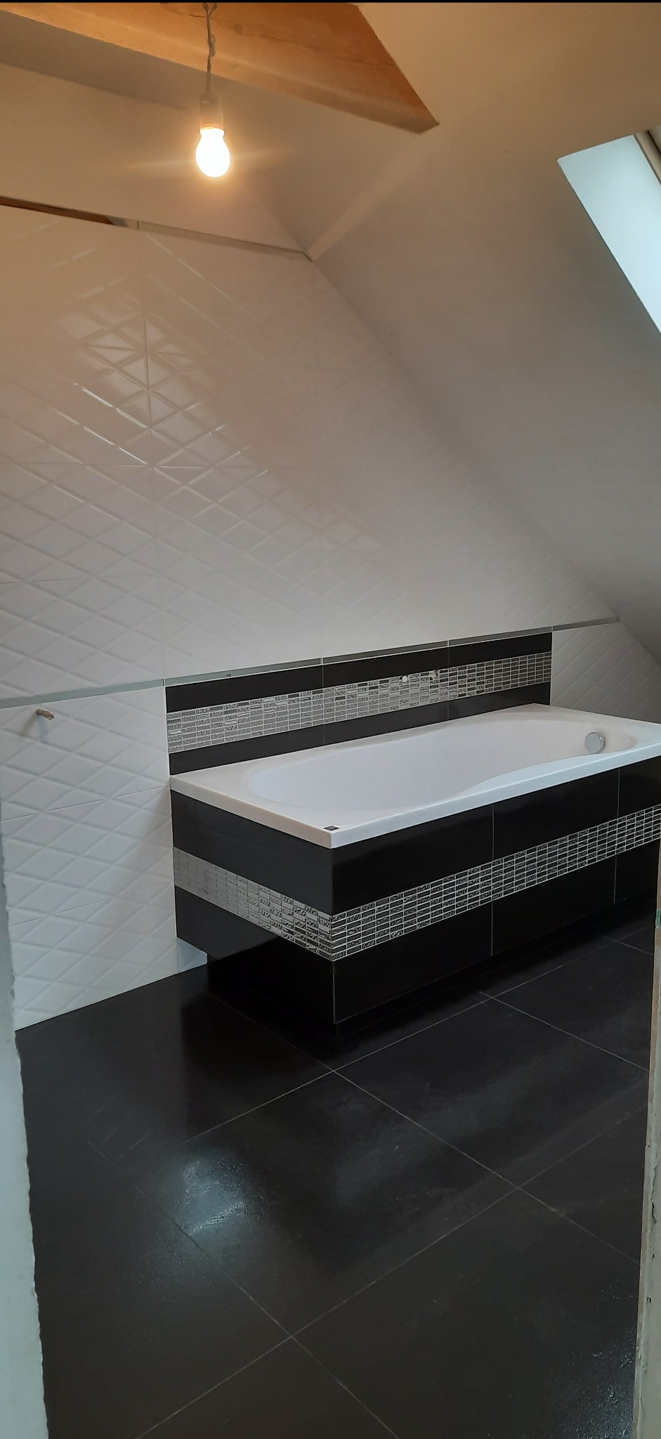 Wykończona łazienka na poddaszu ze skośnym sufitem, białe płytki z geometrycznym wzorem na ścianach, czarne płytki na podłodze i obudowie wanny z mozaikowymi wstawkami.