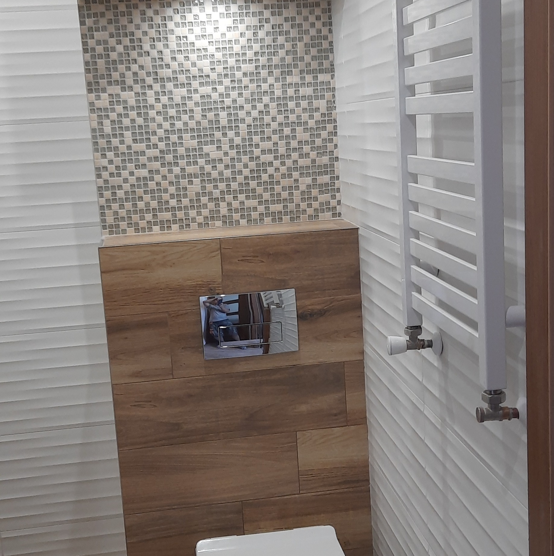 Fragment łazienki z mozaiką, płytkami imitującymi drewno, grzejnikiem drabinkowym i chromowanym przyciskiem spłuczki, odbicie osoby w przycisku.